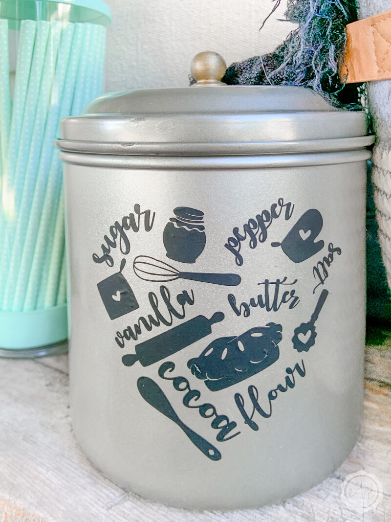 A stainless steel kitchen themed canister with an iron on decal on the front in the shape of a heart. The heart is made up of different baking icons like sugar, pepper, an oven mitt, rolling pin, whisk, pie, butter, vanilla, salt, cocoa, flour and a wooden mixing spoon. 