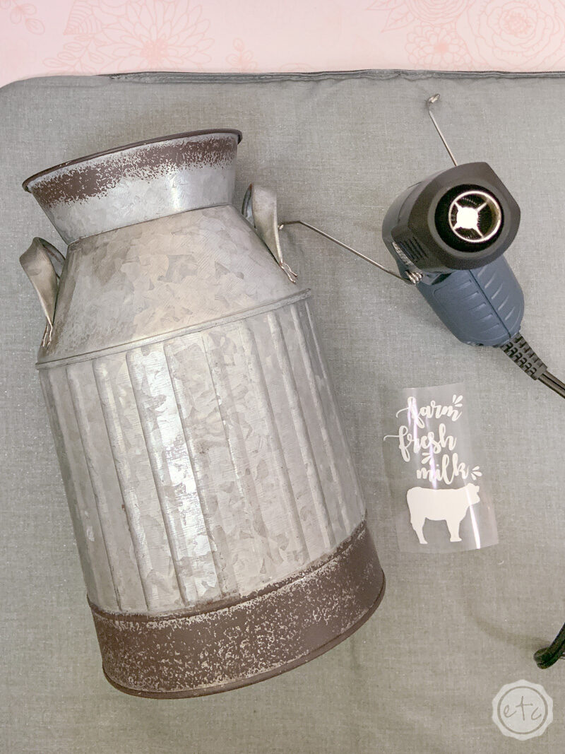 Setting up the work station in order to apply htv to a tin milk jug. A weeded iron on design sits next to the milk jug and wagner heat gun ready to be applied.